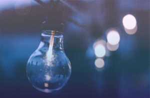 Photo of a large round light bulb glowing in sharp focus in the left foreground with a dark background. To the right, and further back, is a row of several more round glowing lights in soft focus, extending away from the camera