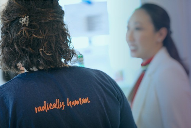 Candid photo of two Promise54 staff members talking. One's back is to the camera, wearing a blue shirt that reads "radically human" in orange letters. The other is out of focus, wearing a white blazer.